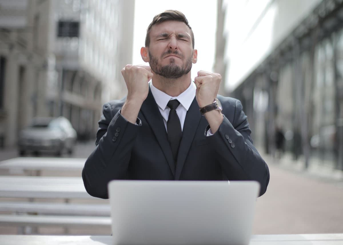 A man in a suit grimacing with clenched fists at a laptop.